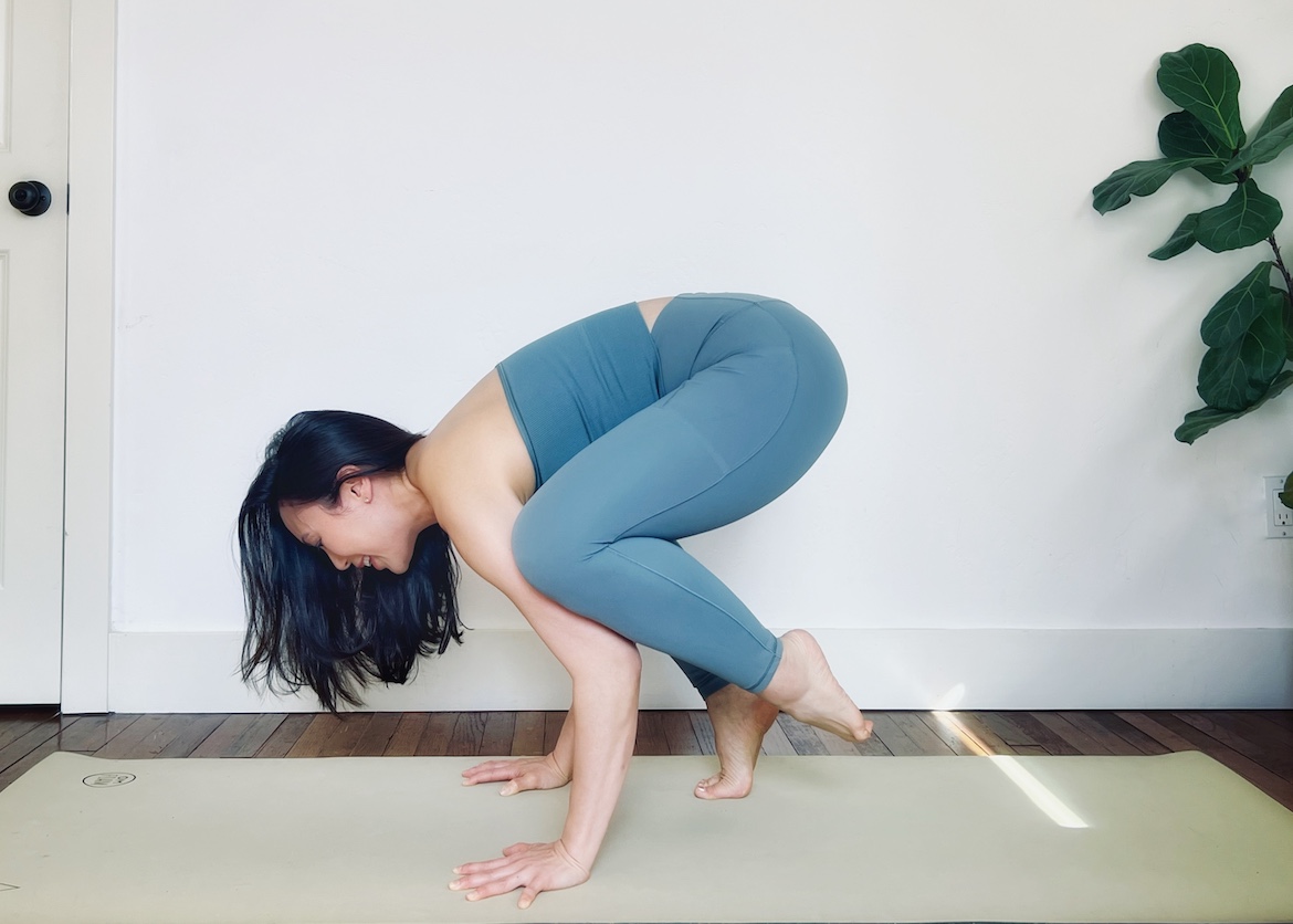 female yoga teacher wearing a matching blue set shows how to do crow pose with one foot on the mat for beginners