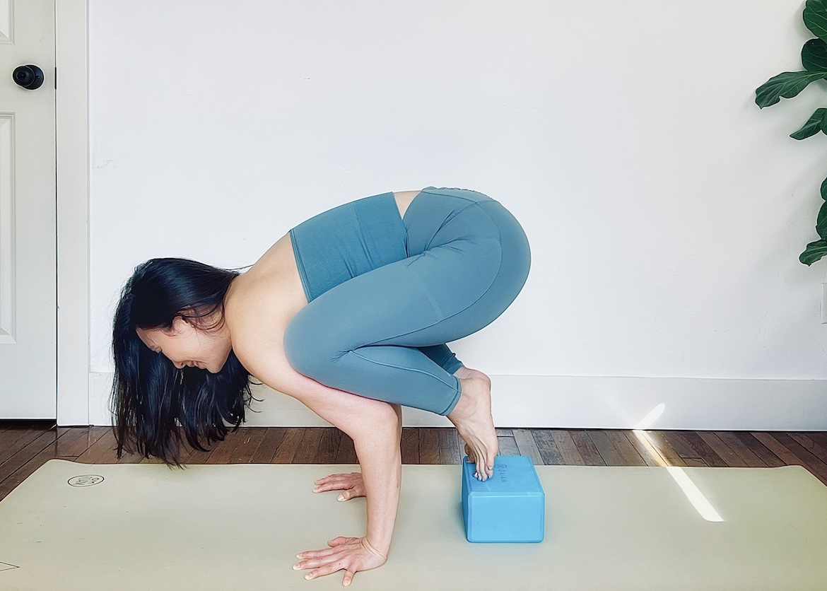 female yoga teacher wearing a matching blue set shows how to do baby crow pose using a block under her feet