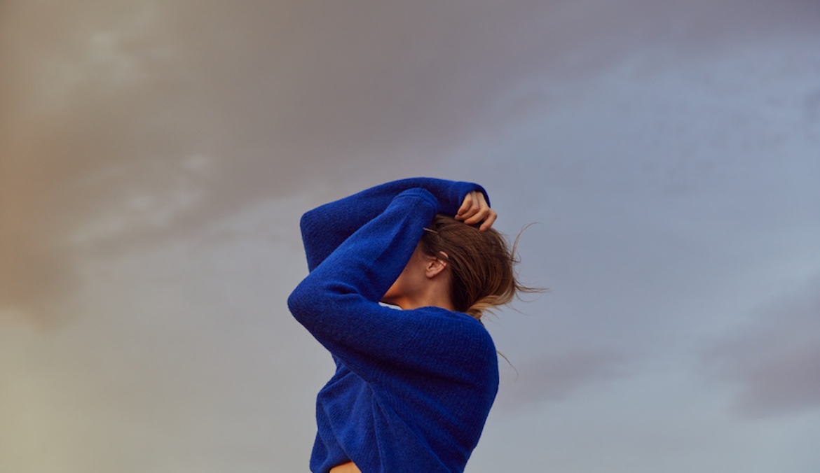 Woman wearing blue sweater covering her face against backdrop of pastel sky