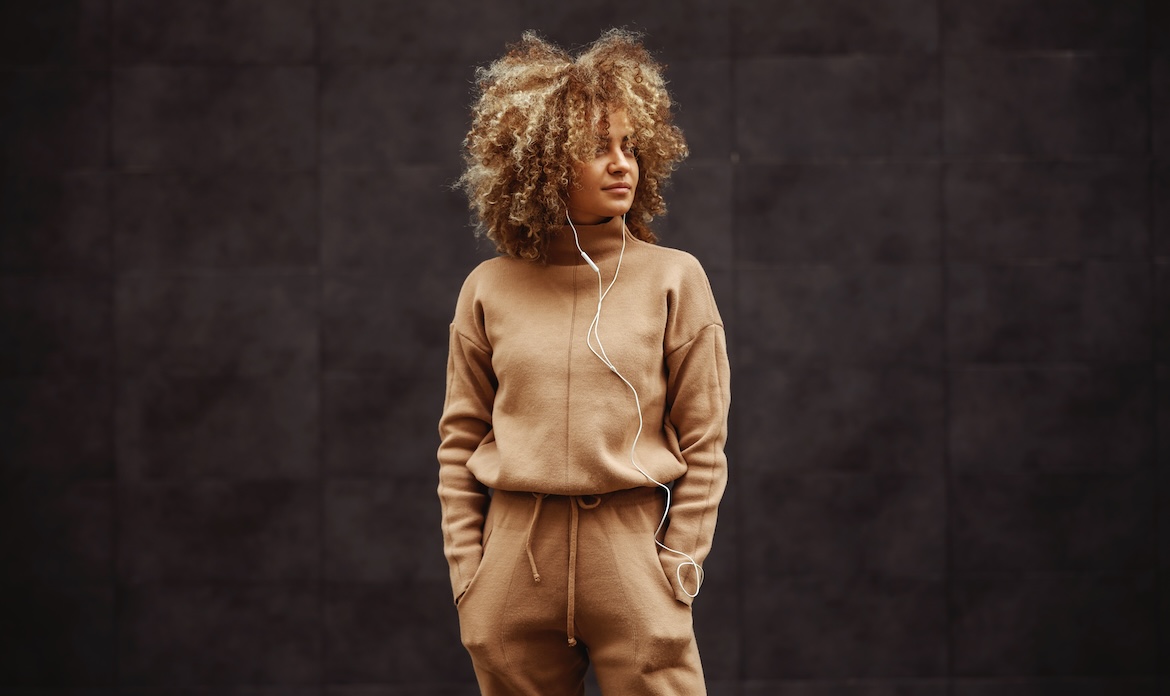 A woman in a sweatsuit stands against a wall listening to music through earphones.