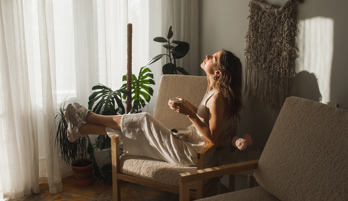 Positive young female in casual clothes smiling while drinking herbal tea during weekend at home. Caucasian girl enjoying a hot cup of tea at living room. Coffee break. Morning mood.