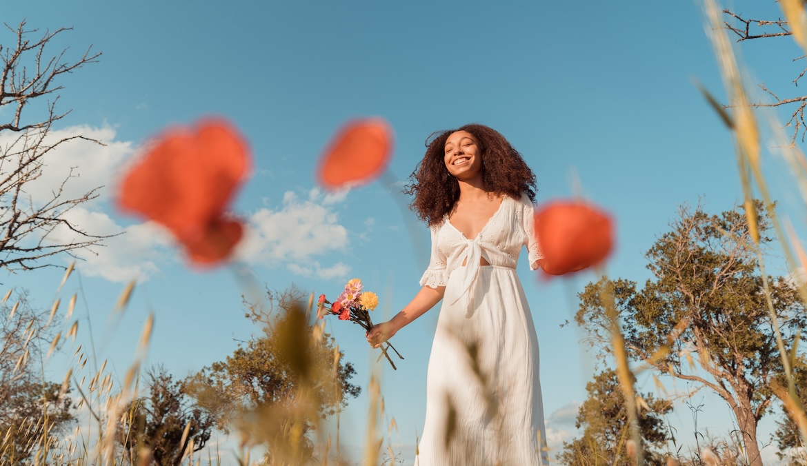 Low angle portrait of a young multiethnic woman with afro hair, wearing a white dress, surrounded by plants and flowers in the countryside and smiling.
