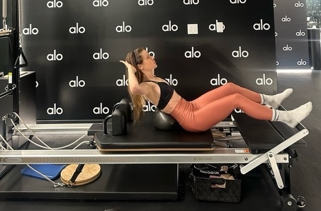pilates instructor does a crunch on the reformer using a pilates ball under her back