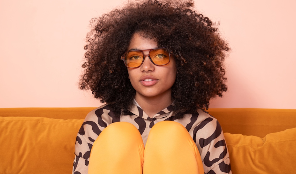 A captivating portrait of a young woman with voluminous curly hair and amber-tinted glasses, seated against a mustard yellow couch, her confident pose and patterned outfit exuding a casual yet sophisticated aura