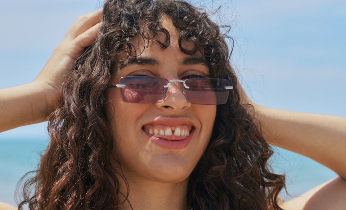 Woman with long curly hair is at the beach in a sunny day, happy and smiling. She is wearing vintage purple tinted sunglasses and grabbing her head. She has a teeth gap.
