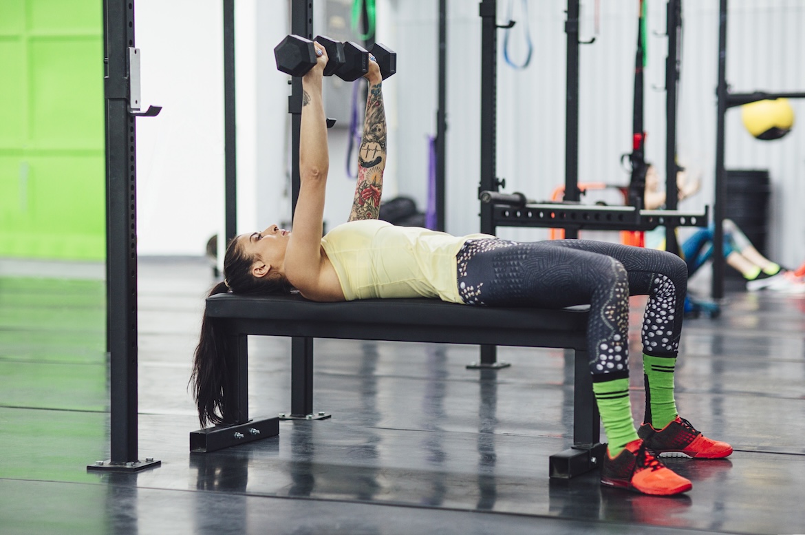 Person wearing yellow tank top and black leggings doing a bench press with dumbbells