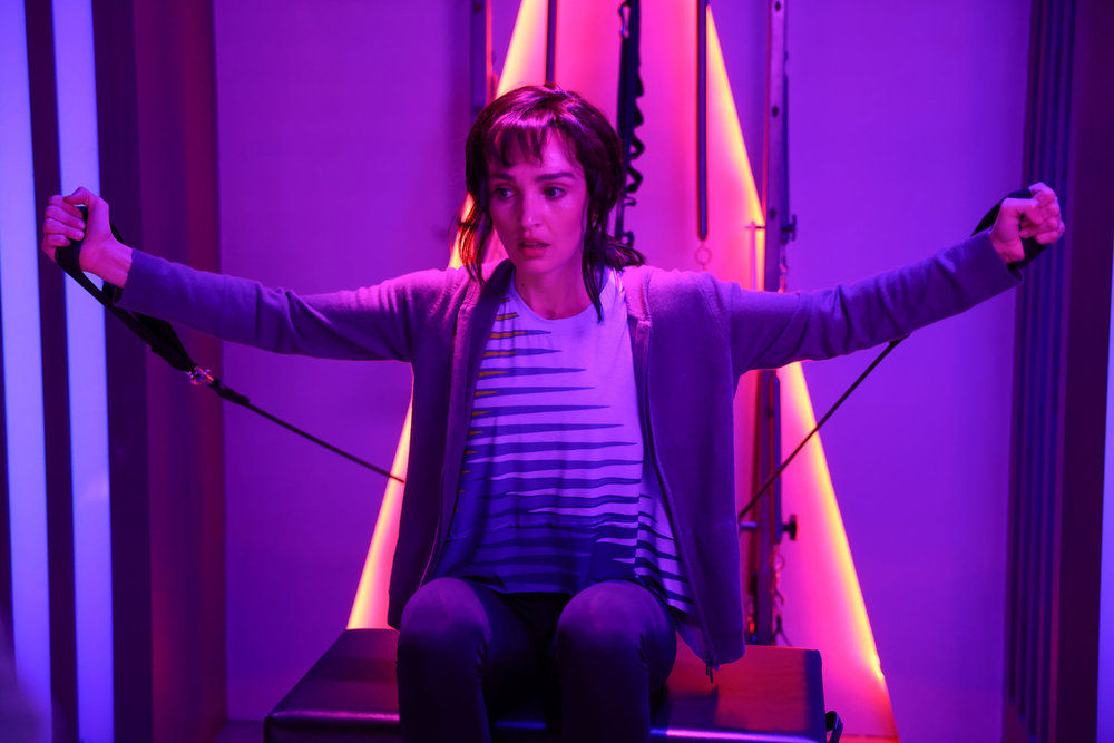 SNL star Chloe Fineman on a pilates machine in a purple and pink neon lit room.