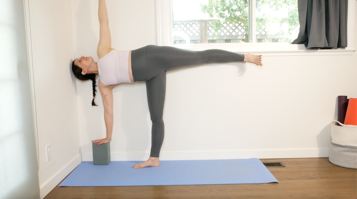 female yoga teacher with her dark hair in a braid demonstrates how to use a wall to modify half moon, using a block on her lower hand, standing on one leg, and lifting the other hand to the sky