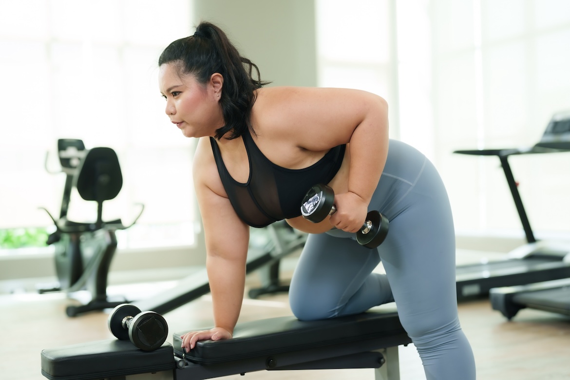 Person wearing black sports bra and blue leggings doing triceps workout in gym with dumbbells